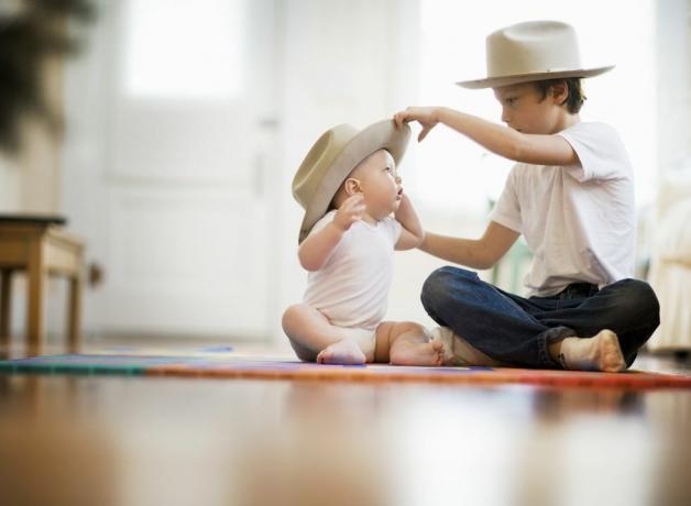 selektywny widok ostrości dwóch braci, jeden ma dziewięć lat, jeden ma osiem miesięcy, para siedzi ze skrzyżowanymi nogami na podłodze starszy chłopiec zakłada pasujący kapelusz na głowę dziecka