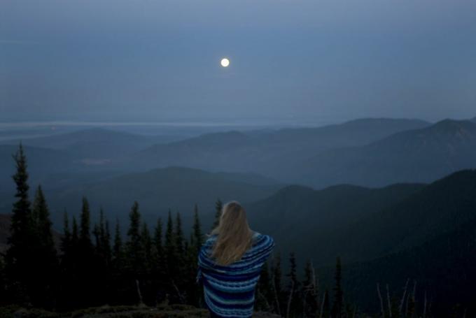 kobieta owinięta w koc patrząca na górzysty krajobraz z pełnią księżyca, widok z tyłu
