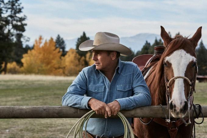 kevin costner w yellowstone obok konia opartego o płot z liną w dłoniach, ubrany w wyblakłą niebieską dżinsową koszulę i beżowy kowbojski kapelusz