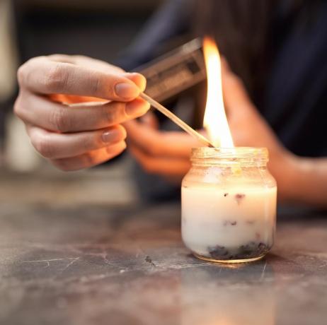 przytnij anonimową kobietę z płonącą zapałką zapalającą aromatyczną świecę w szklanym słoju umieszczonym na marmurowym stole w domu