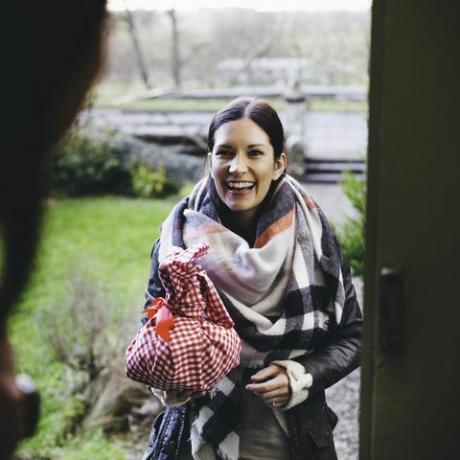 kobieta uśmiecha się stojąc na progu wchodząc do koleżanki owinięta w ciepłe ubranie i trzyma prezent owinięty w czerwono-białą kratkę