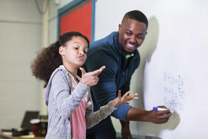 wielorasowa 9-letnia dziewczynka na lekcjach matematyki, stojąca przy tablicy ze swoim nauczycielem, młody Afroamerykanin w swoim 20. Nauczyciel patrzy na swojego ucznia ze szkoły podstawowej i czeka, aż ona będzie próbowała znaleźć odpowiedź, skupiając się na dziewczyna